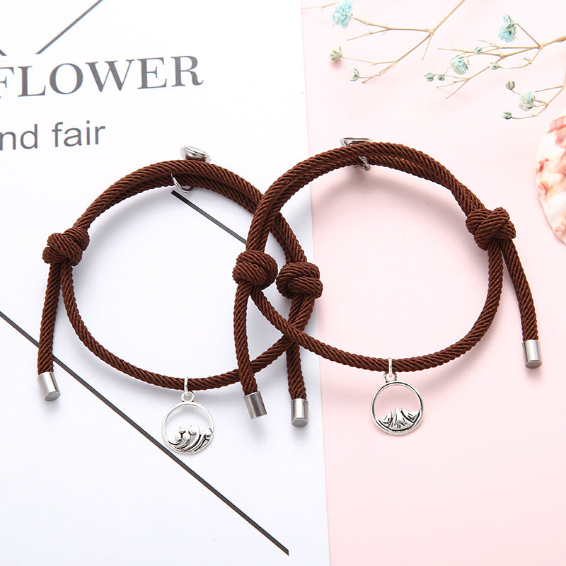 Two brown cord New Alloy Eachother Couple Bracelet with silver charms on a white and pink background.